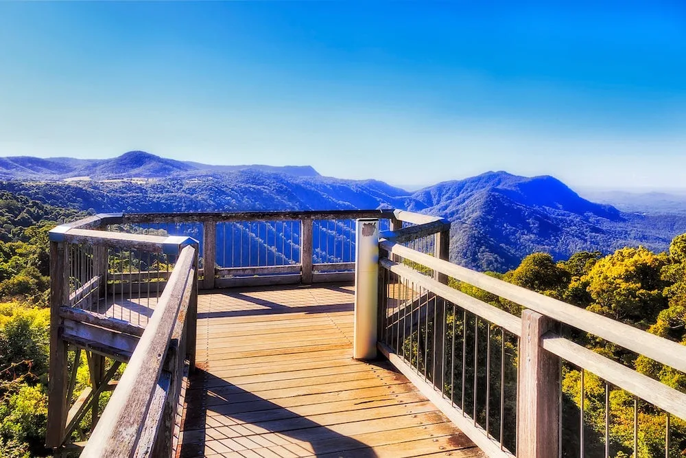 A wooden lookout deck with metal railings offers a panoramic view of rolling blue mountains and lush green forests