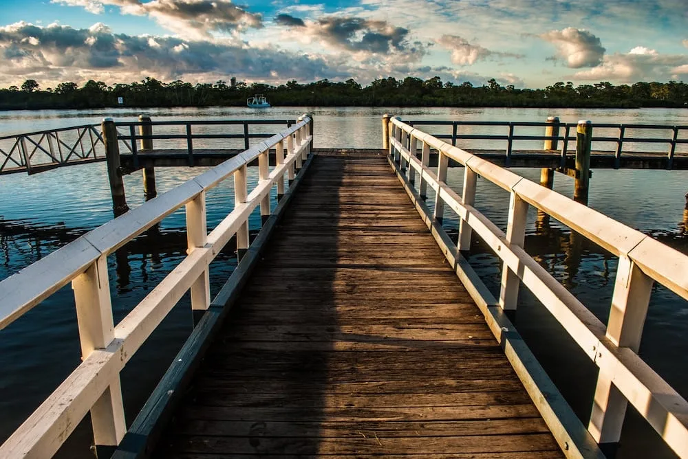 A wooden pier with white railings extends toward a calm river under a cloudy sky