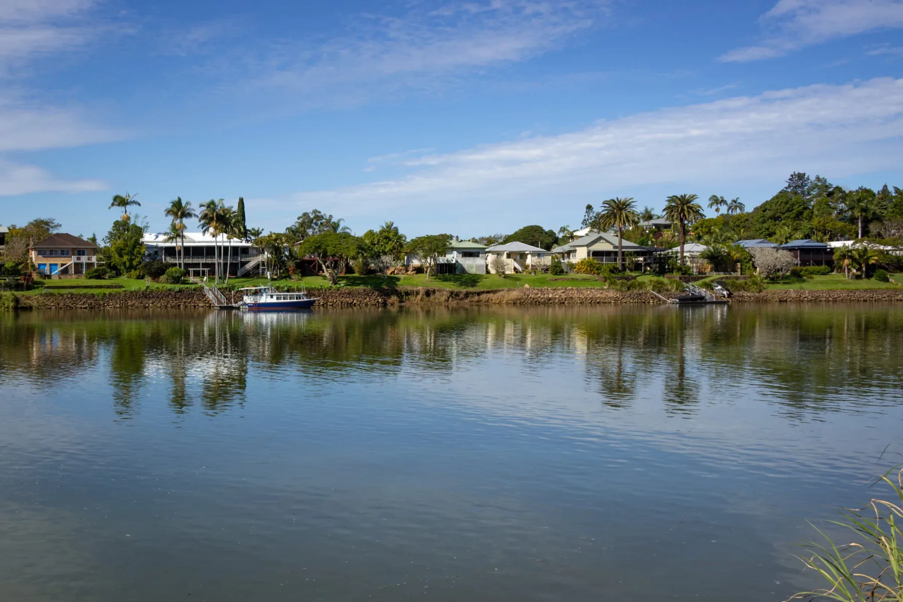 A row of waterfront houses and palm trees reflecting in a calm river under a blue sky