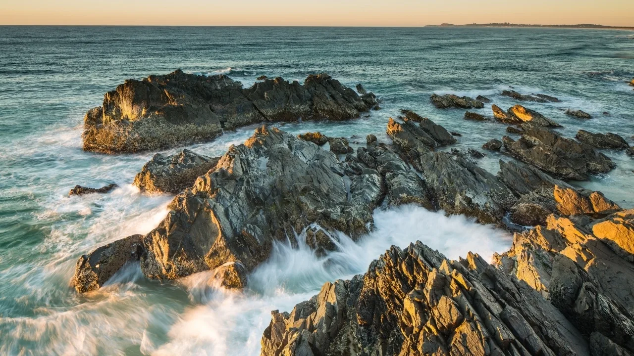 A long-exposure shot of ocean waves crashing against jagged, dark coastal rocks.
