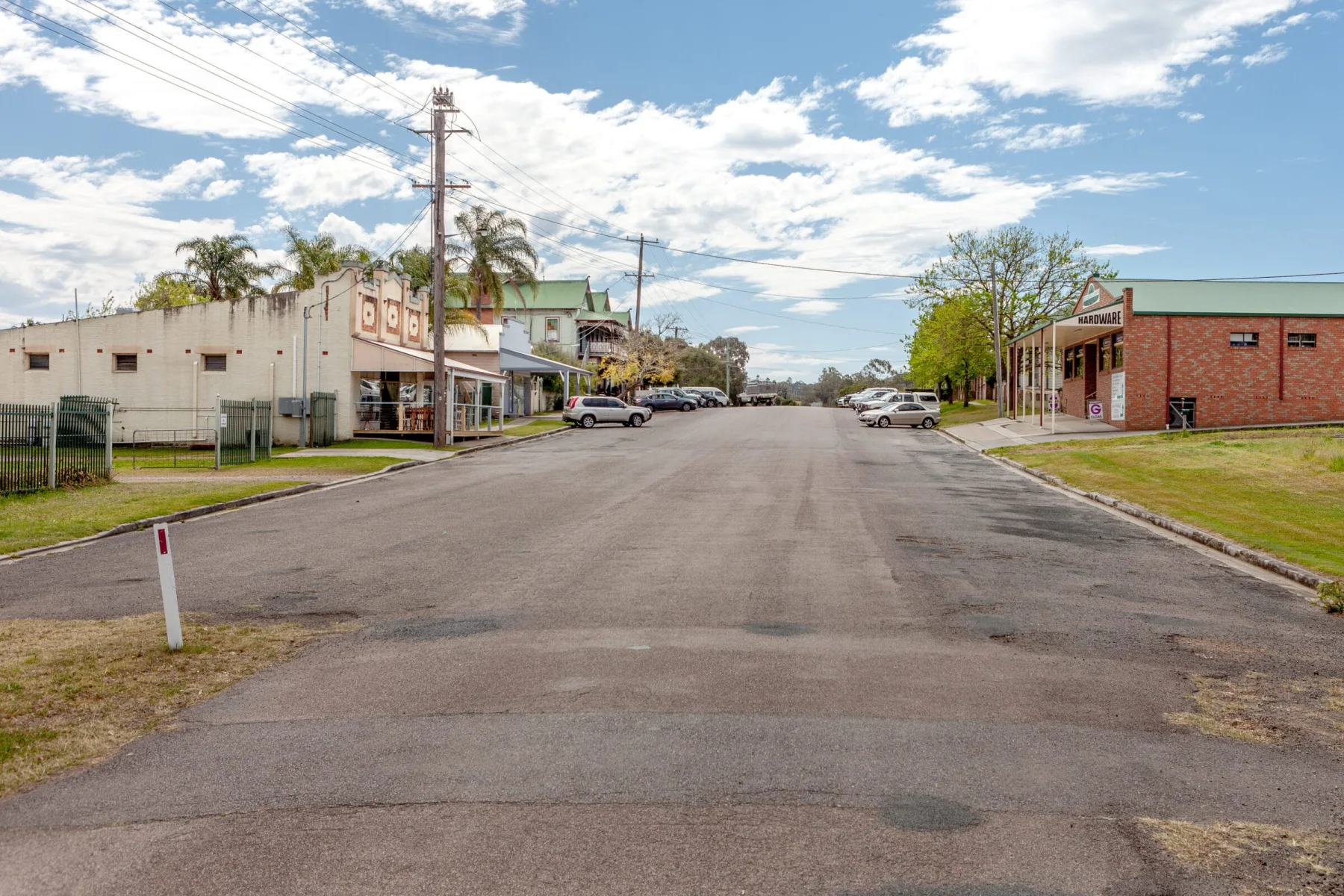 A wide, quiet street in a small town lined with buildings, including a brick hardware store on the right, under a blue sky with scattered clouds.