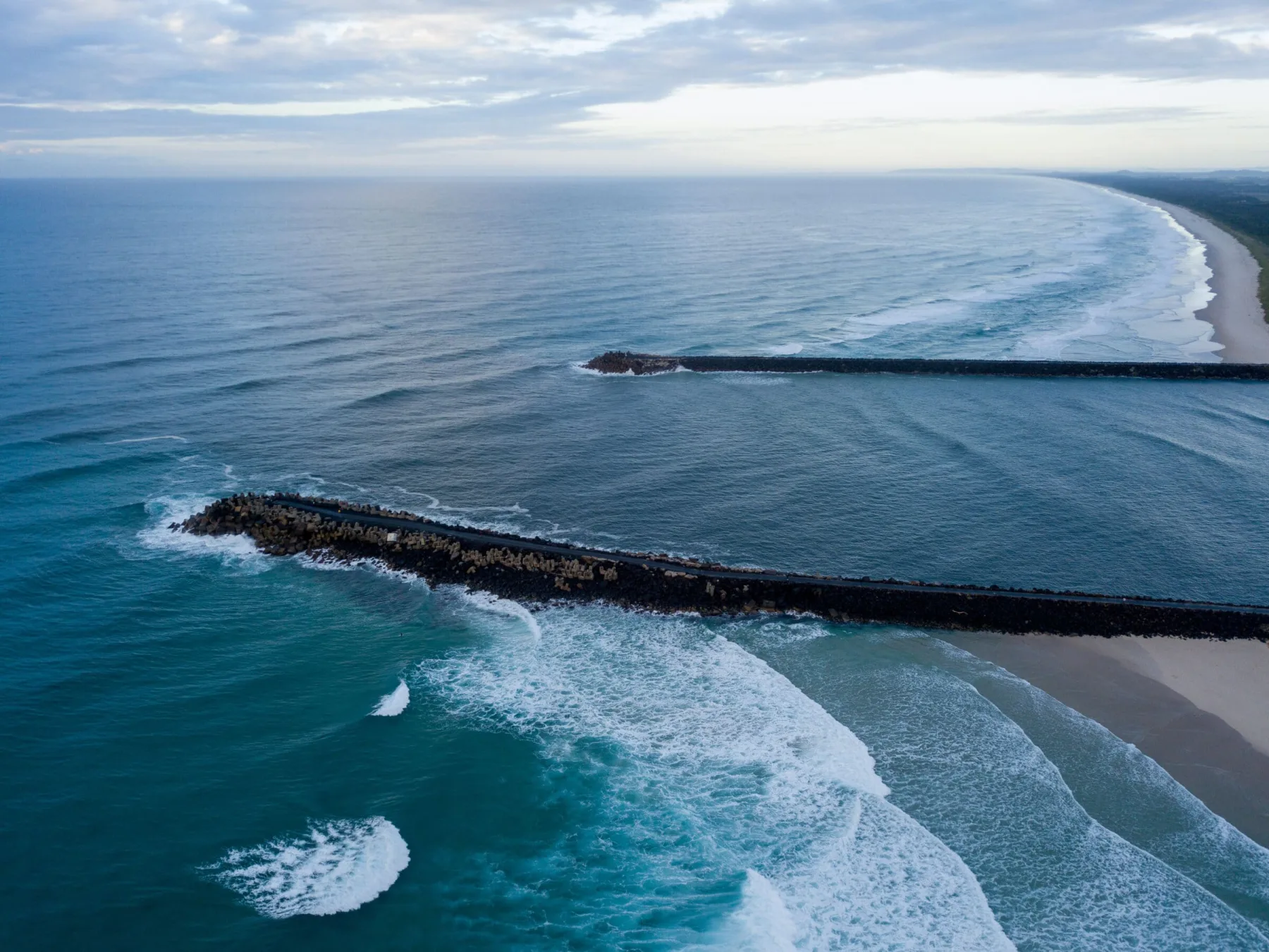 An aerial view looking out at two long rock breakwaters extending into a vast blue ocean