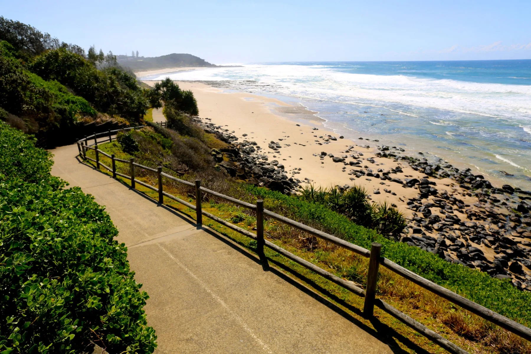 A paved coastal walkway with a wooden fence winds along a lush hillside, overlooking a wide sandy beach scattered with dark rocks.