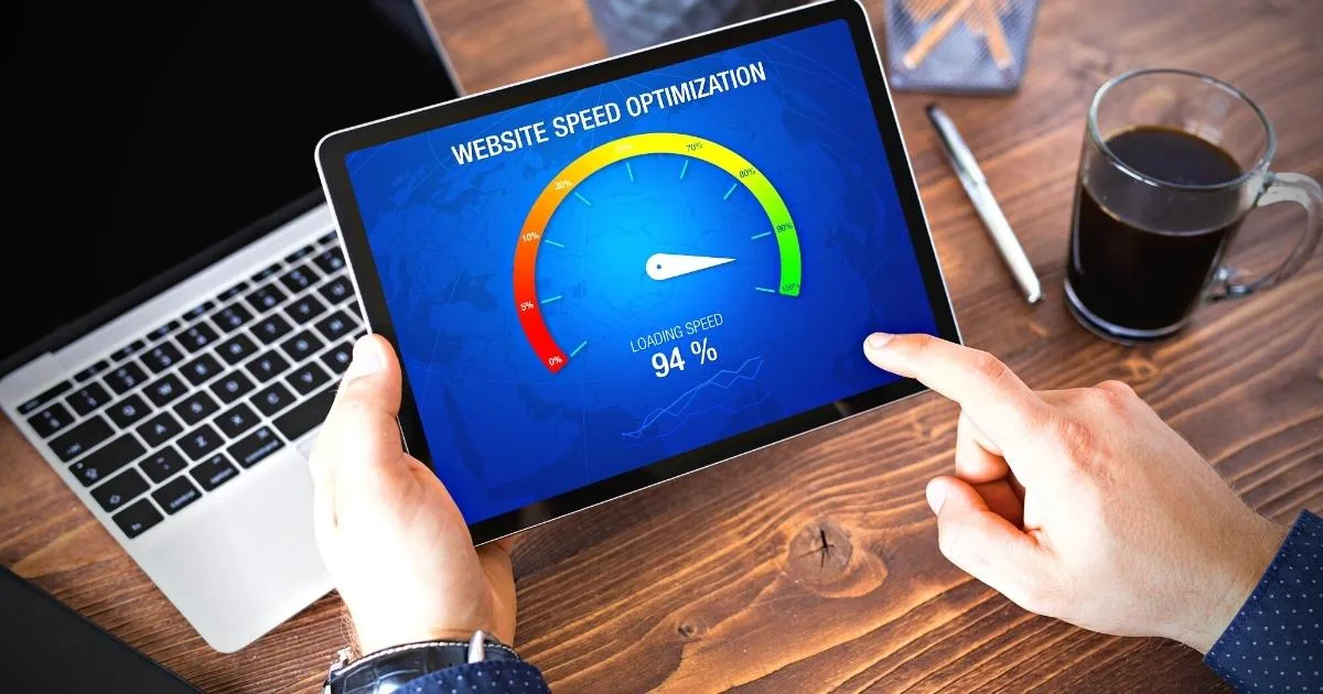 A person holds a tablet over a wooden desk, displaying a colourful speedometer graphic labelled 'WEBSITE SPEED OPTIMIZATION.