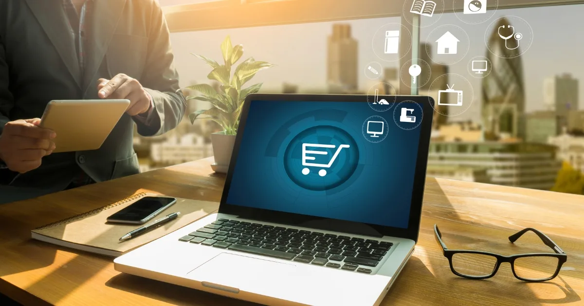 A man works at a wooden desk with a city skyline in the background, using a tablet while a laptop screen displays a glowing shopping cart icon.