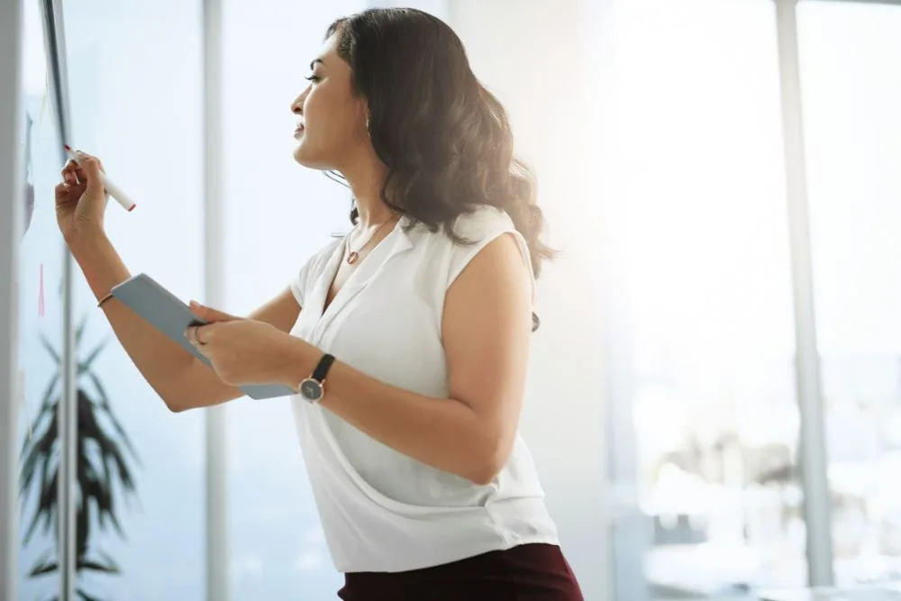 A profile view of a professional woman in a white blouse writing on a glass whiteboard with a marker while holding a tablet.