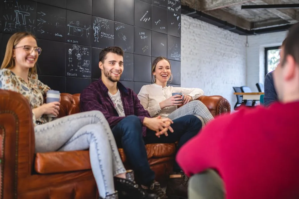 A group of three smiling young professionals sitting on a brown leather tufted sofa in a modern, casual office setting.