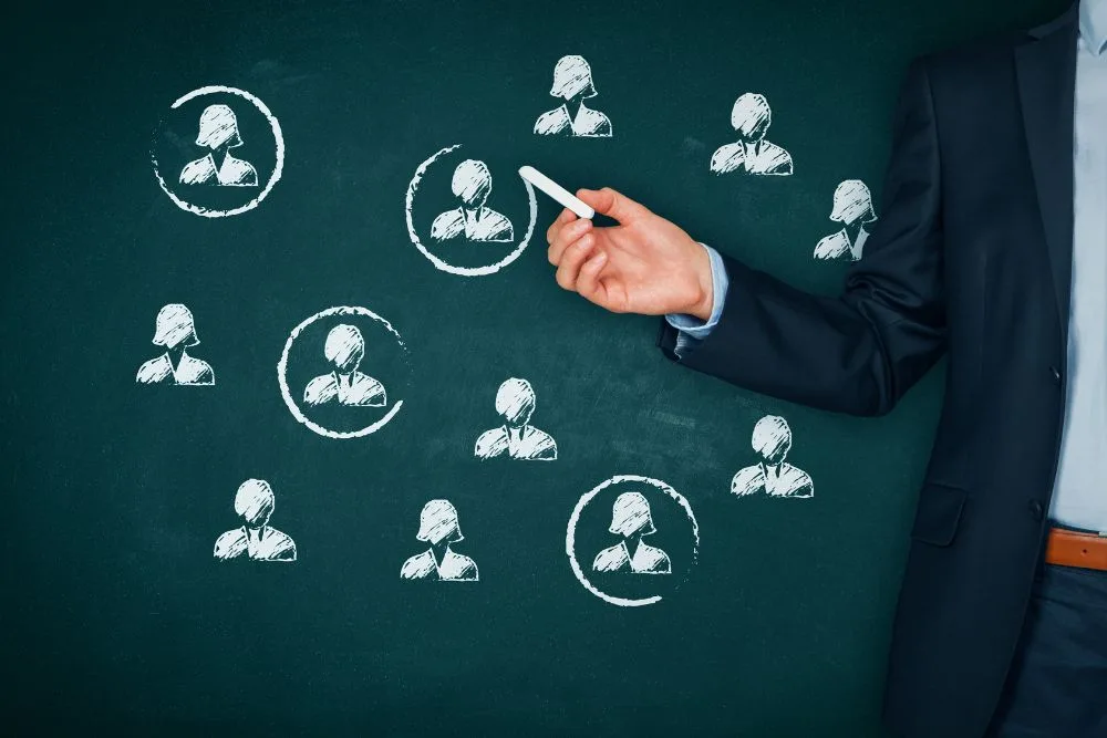 A person in a dark business suit uses a piece of white chalk to draw a circle around one of several human silhouettes sketched on a dark green chalkboard.
