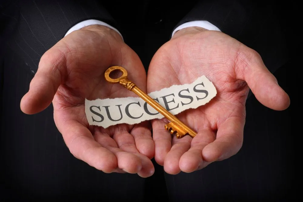 A close-up shot of a person in a business suit holding their cupped hands open, revealing a small, vintage-style golden key resting on a torn piece of paper.