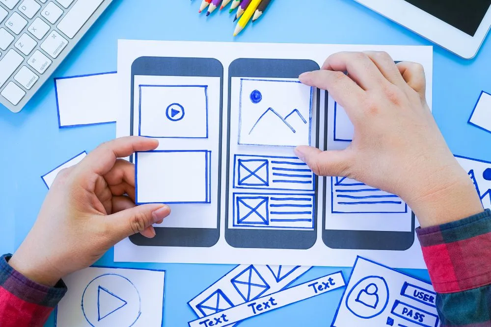 An overhead shot shows a designer's hands arranging paper cutouts on a hand-drawn mobile app wireframe.