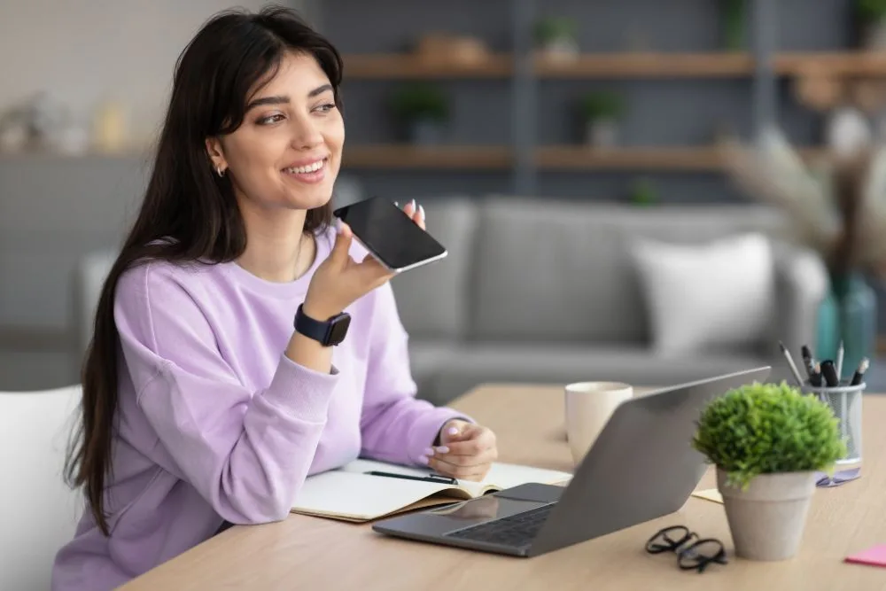 A smiling young woman in a lavender sweatshirt sits at a wooden desk