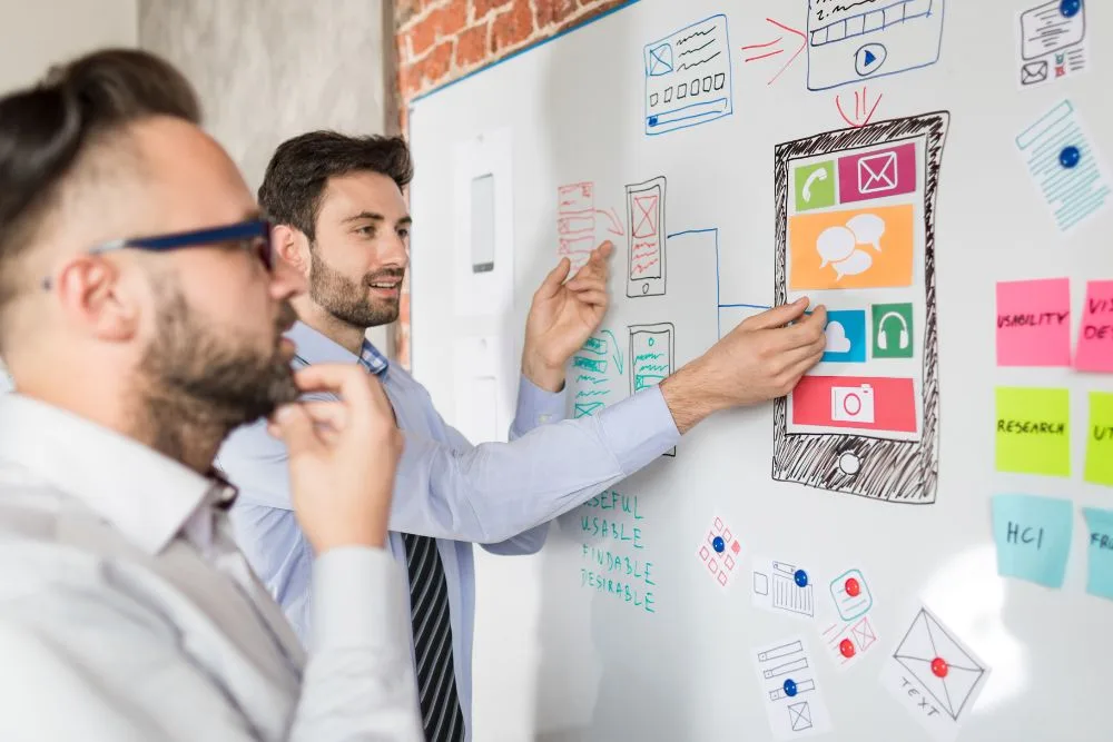 Two men in a professional setting collaborate on a mobile app design using a large whiteboard.