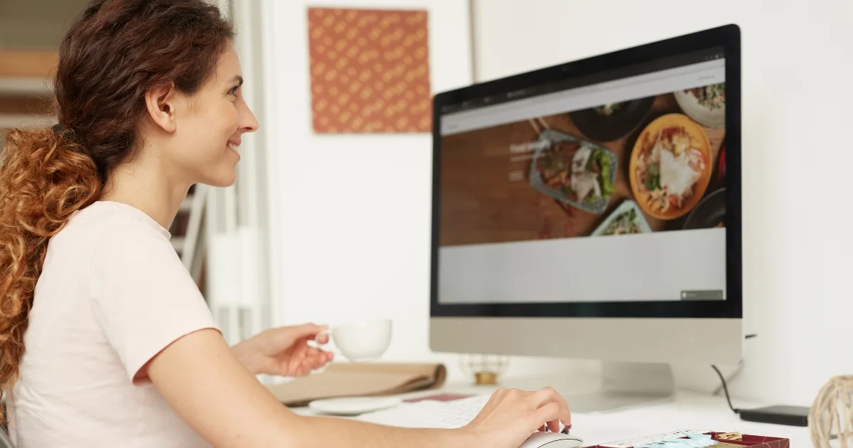 A woman with curly hair smiling and holding a cup while looking at a desktop computer screen