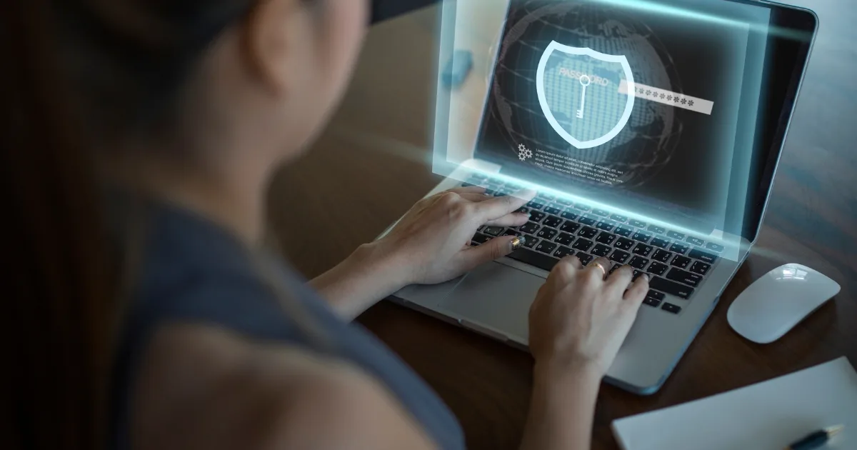 An over-the-shoulder view of a person typing on a laptop that features a glowing digital overlay of a shield