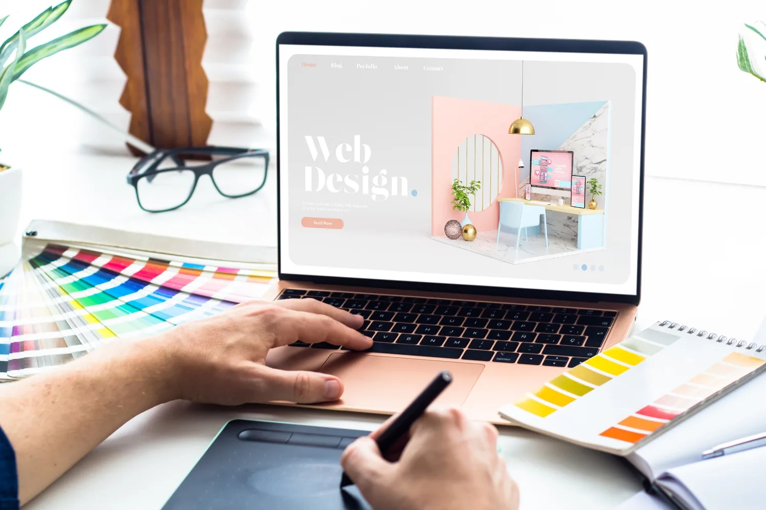 A designer’s hands working at a bright, white desk, using a laptop and a digital drawing tablet with a stylus.