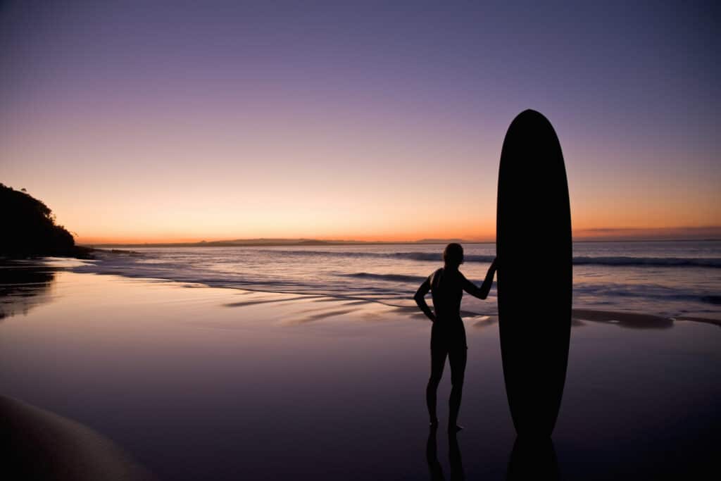 A silhouette of a lone surfer standing on a wet, reflective beach at twilight
