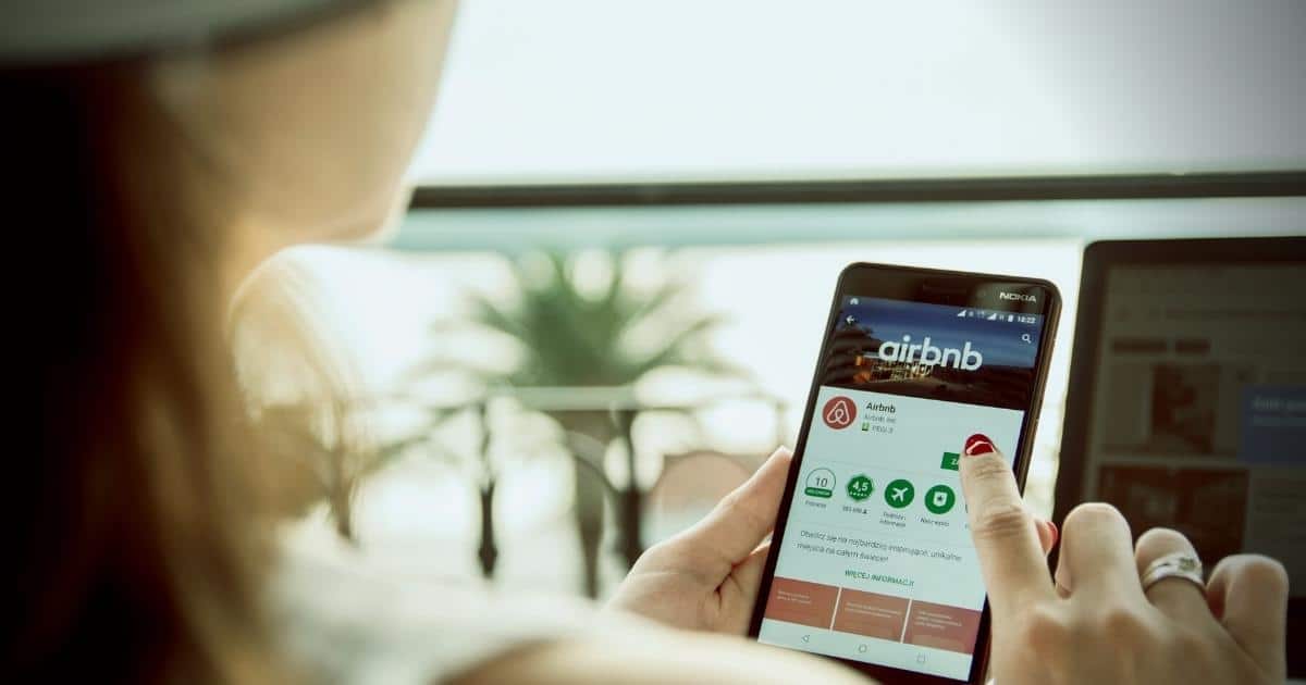 An over-the-shoulder view shows a person holding a smartphone and navigating the Airbnb mobile app in an app store.