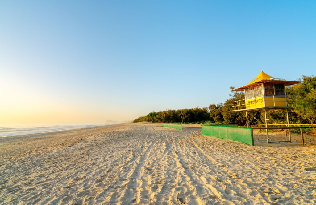 A wide, low-angle shot of a tranquil beach at dawn features a yellow lifeguard tower standing on the right, surrounded by a line of coastal trees.