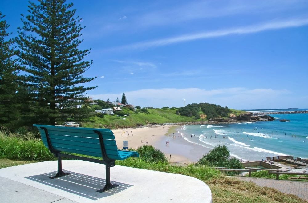 A teal park bench sits on a concrete lookout, offering a high-angle view of a sandy beach filled with people